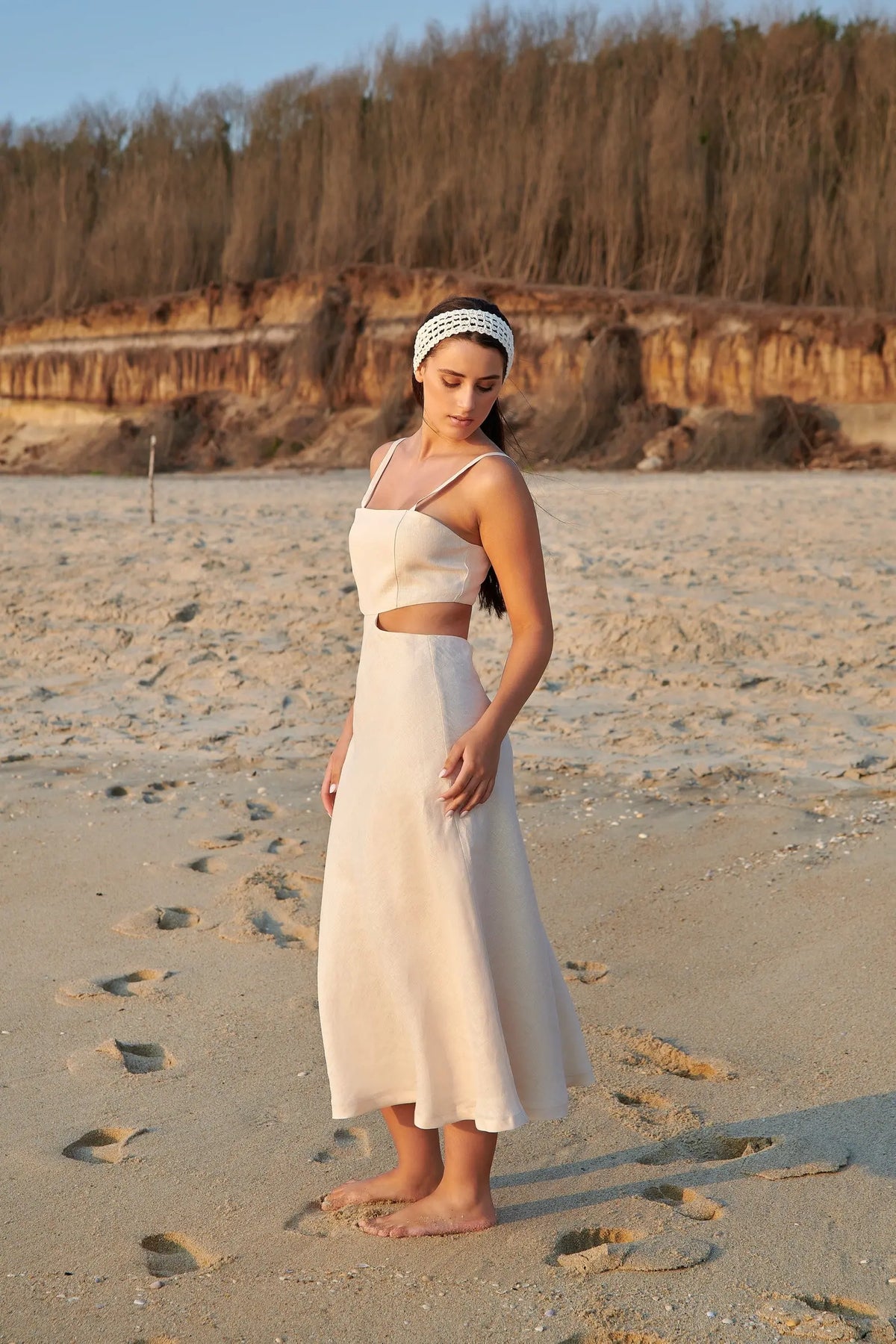 Vue de profil de la robe Maya longue en lin beige, portée sur une plage, montrant sa taille découpée et sa jupe fluide.