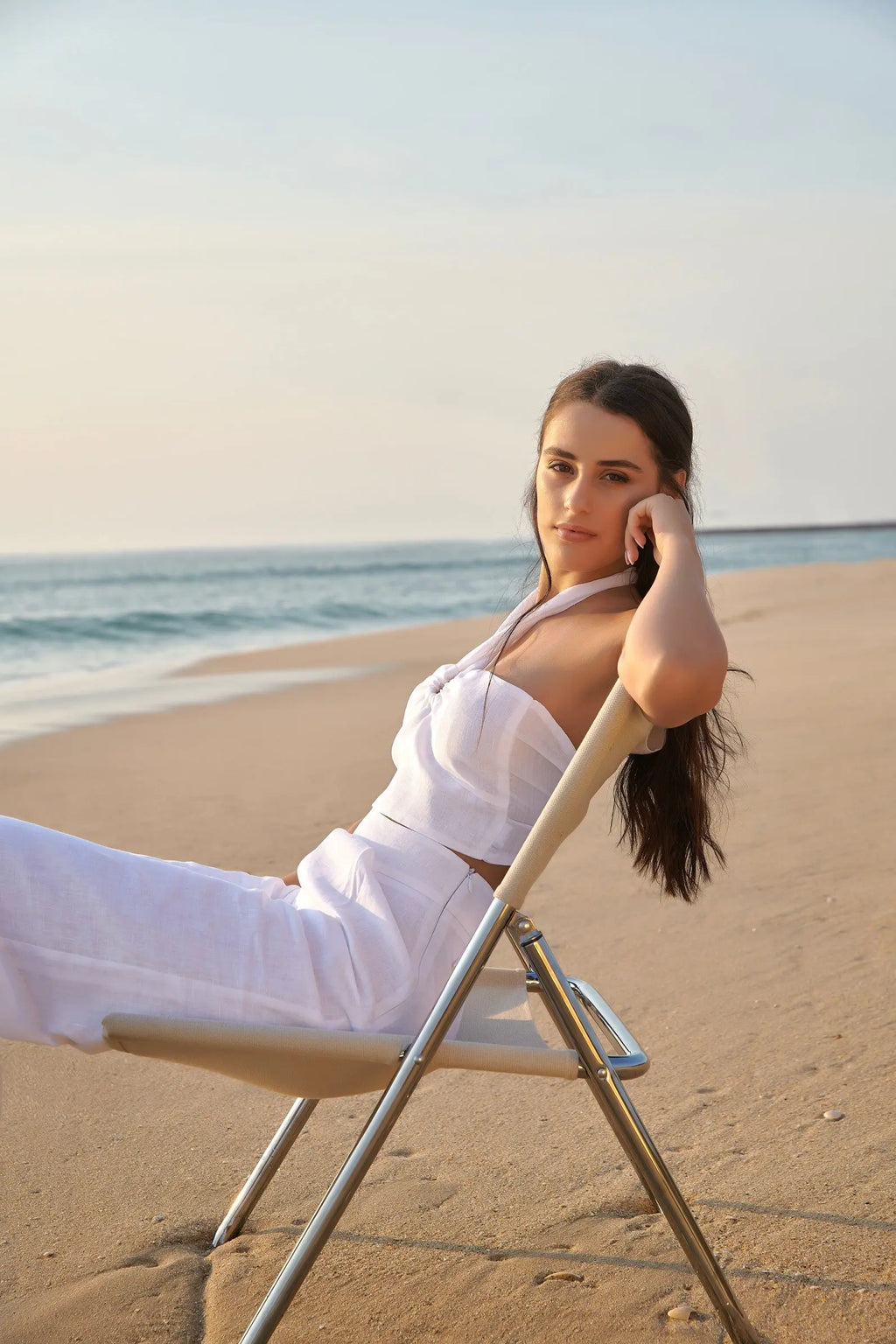 Femme assise sur une chaise longue au bord de la mer, portant le top Léa blanc en lin et un pantalon blanc.
